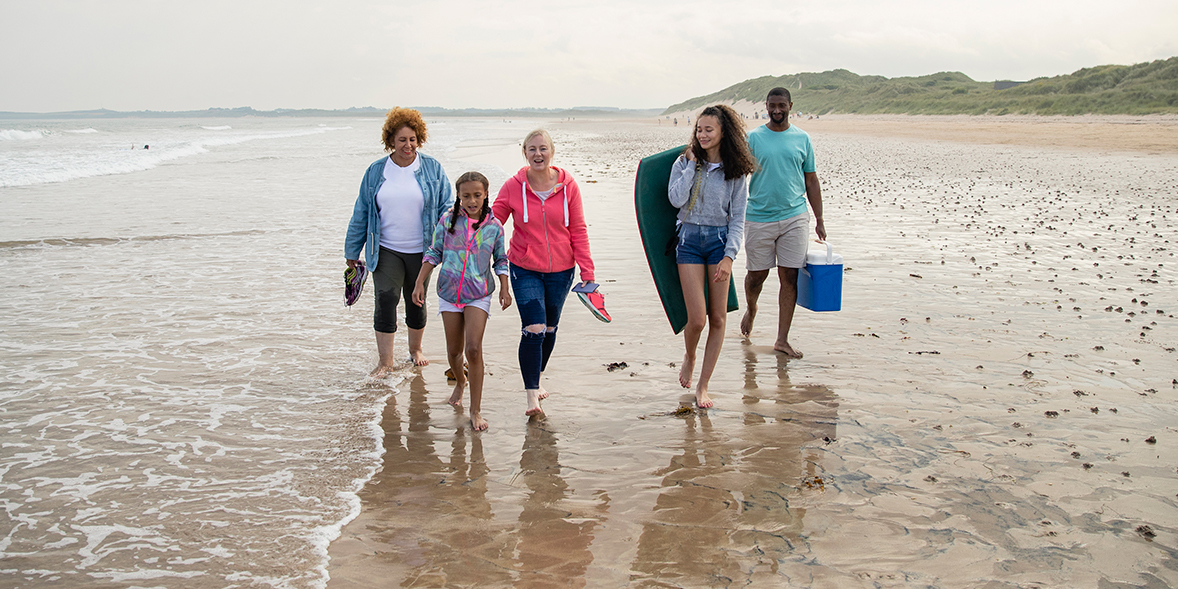 Family walking barefoot on beach, one person holding a blue cool box